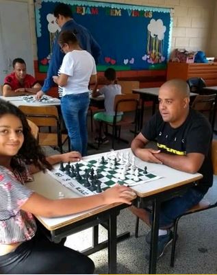 Foto na qual  aparece o autor (instrutor de xadrez), alunos com seus pais em oficina para aprender jogar xadrez. A frente e a esquerda da sala apresenta o professor orientando um pai sentado e a filha filha em pé e ao fundo e mais a esquerda um pai calvo e a filha sorridente sentada à mesa olhando para o fotógrafo.