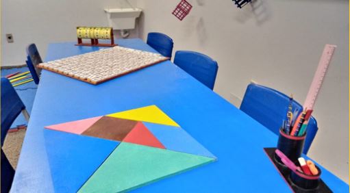 Foto da Sala de Recursos Maker apresentando uma mesa azul e 5 cadeiras também na cor azul. Em cima da mesa tem três jogos: Roleta das operações matemáticas, Caça-palavras e o  Tangram. Todos tamanho ampliado. Tem também um porta-canetas na mesa.

