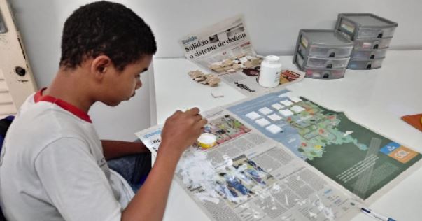 Foto de um estudante, uniformizado pintando as peças de um jogo. A mesa está coberta com jornal.


