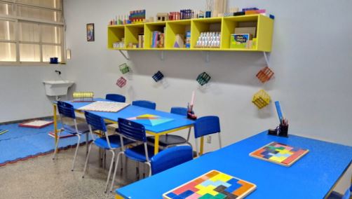 Foto da Sala de Recursos Maker. Acima e à direita está uma estante amarela onde estão guardados alguns jogos. Logo à direita do armário está um quadro branco. 
À esquerda e abaixo tem uma mesa azul e 6 cadeiras também na cor azul. Logo à direita está outra mesa azul com 4 cadeiras na cor azul. Em cima das mesas têm jogos. 
