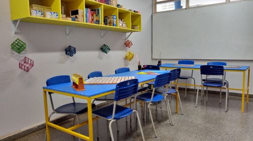 Foto da Sala de Recursos Maker. Acima e à direita está uma estante amarela onde estão guardados alguns jogos. Logo à direita do armário está um quadro branco. 
À esquerda e abaixo tem uma mesa azul e 6 cadeiras também na cor azul. Logo à direita está outra mesa azul com 4 cadeiras na cor azul. Em cima das mesas têm jogos. 
