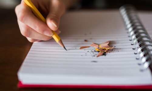 Imagem - representa o ato de escrever - temos: um caderno aberto e sobre ele uma mão segura um lápis.