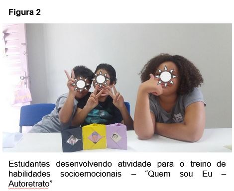 Descrição da Imagem: Três crianças sentadas, sorrindo e fazendo gestos de confirmação da realização da atividade proposta, a construção de um autorretrato.
