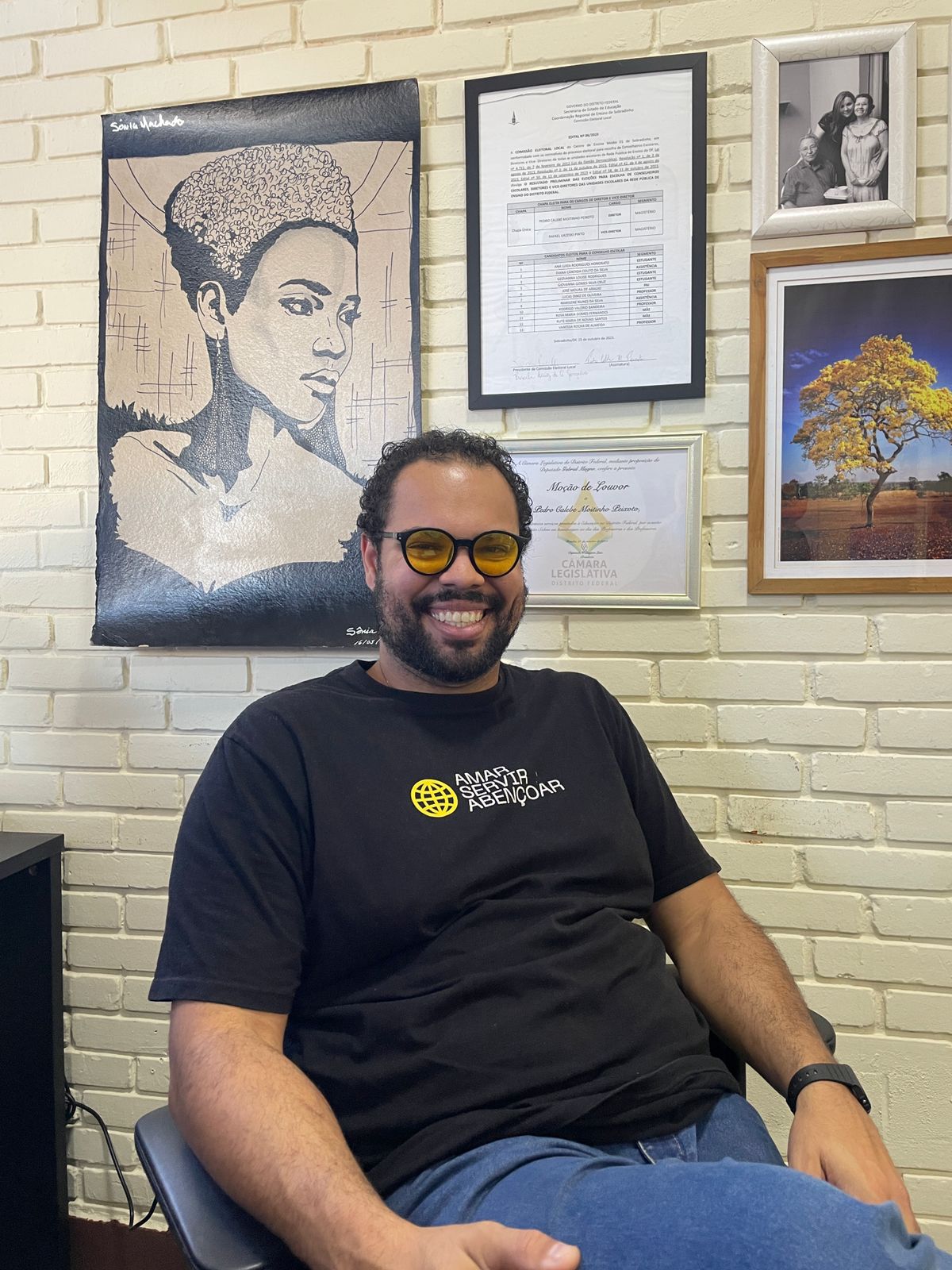 Audiodescrição: Pedro Calebe Moitinho Peixoto, homem pardo de aproximadamente 35 anos, está sentado em cadeira giratória azul, com postura relaxada e sorriso amplo. Tem barba curta e cabelos crespos. Usa camiseta preta com a inscrição 'Amar, Servir, Abençoar' em letras amarelas, calça jeans e óculos com lentes levemente amareladas. No pulso esquerdo, um relógio escuro. Ao fundo, parede de tijolos brancos com quadros artísticos emoldurados, um edital de direção com seu nome visível e uma Moção de Louvor da Câmara Legislativa do Distrito Federal.