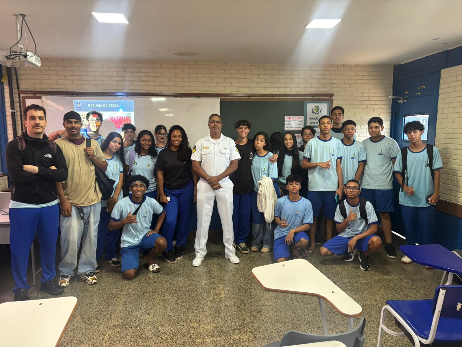Audiodescrição: Foto tirada no interior de uma sala de aula de escola pública. O ambiente tem paredes de tijolos à vista brancos, piso de granilite bege e iluminação fluorescente no teto. Um grupo de cerca de vinte jovens estudantes está reunido em pé no centro da sala, posando para a foto junto a um homem adulto negro, de uniforme branco da Marinha do Brasil, posicionado no meio do grupo. Os alunos vestem o uniforme escolar azul-claro com logo do GDF. Ao fundo, uma lousa verde e um projetor exibindo uma apresentação com o texto 'Marinha do Brasil'. Há carteiras escolares azuis espalhadas pela sala. A cena retrata uma atividade de orientação profissional com representante das Forças Armadas.