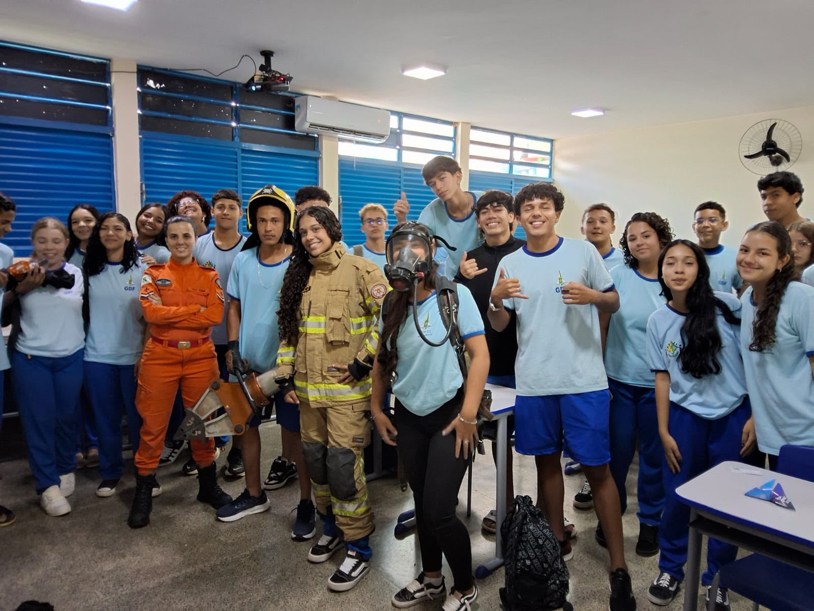 Audiodescrição: Foto em sala de aula do CEM 01 de Sobradinho. O ambiente tem paredes claras, janelas com persianas azuis ao fundo e equipamentos escolares como projetor e ventilador de parede. Um grupo de cerca de vinte e cinco estudantes adolescentes em uniforme azul-claro do GDF sorri para a câmera. Dois jovens se destacam ao centro: uma estudante negra usando o equipamento completo de bombeiro — roupa de combate a incêndio bege com faixas refletivas e capacete amarelo — e outra estudante com uma máscara de respiração industrial sobre o rosto. Ao lado deles, uma bombeira profissional adulta, com uniforme laranja. A cena representa uma atividade prática de educação para segurança e orientação profissional, com alta energia e entusiasmo dos participantes.