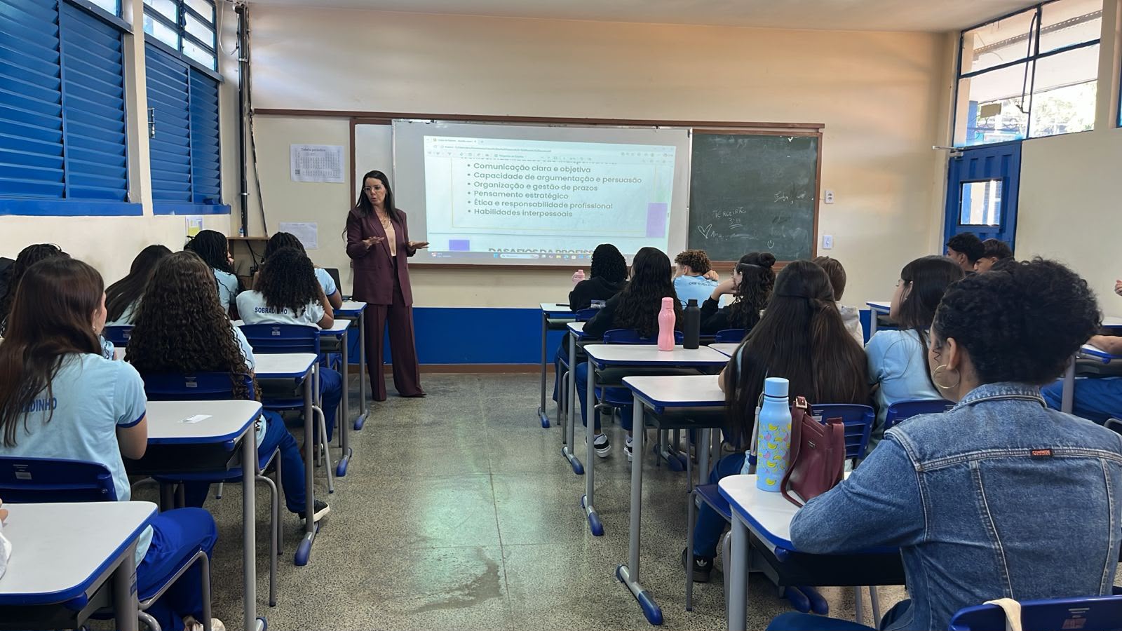 Audiodescrição: Vista interna de uma sala de aula ampla do CEM 01 de Sobradinho, fotografada do fundo para a frente. As janelas à esquerda têm venezianas azuis abertas, deixando entrar luz natural. As carteiras e cadeiras azuis estão organizadas em fileiras, ocupadas por estudantes adolescentes — maioria meninas — de costas para a câmera, usando o uniforme escolar azul-claro. Ao fundo, uma mulher adulta de terno vinho está em pé ao lado de um projetor, conduzindo uma apresentação. Na tela, é possível ler tópicos como 'Comunicação clara e objetiva', 'Capacidade de argumentação e persuasão', 'Organização e gestão de prazos', 'Pensamento estratégico', 'Ética e responsabilidade profissional' e 'Habilidades interpessoais'. Há também uma lousa verde ao fundo. A cena representa uma palestra ou aula sobre habilidades profissionais para os alunos do ensino médio.