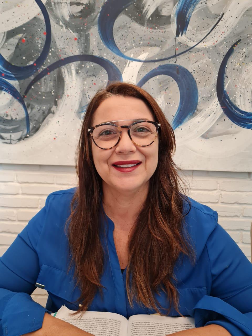 Audiodescrição: Luciana Reis Pereira, mulher branca de meia-idade, sorri de frente para a câmera. Tem cabelos longos, castanho-avermelhados, soltos sobre os ombros. Usa óculos de armaoão arredondada em tons de marrom e dourado, e uma blusa azul royal de mangas compridas. Está sentada à frente de um livro aberto, com os braços apoiados sobre a mesa. Ao fundo, uma parede de tijolos brancos e uma tela abstrata grande, com pinceladas em azul marinho, cinza e branco, decorada com respingos coloridos. A expressão é acolhedora e sorridente.