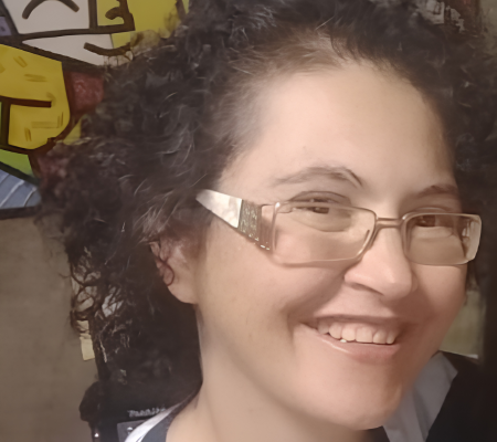 Foto de Fatima Ali Abdalah Abdel Cader-Nascimento: mulher de cabelos cacheados escuros, usando óculos de armação escura, sorriso aberto, vestindo blusa estampada e colorida.