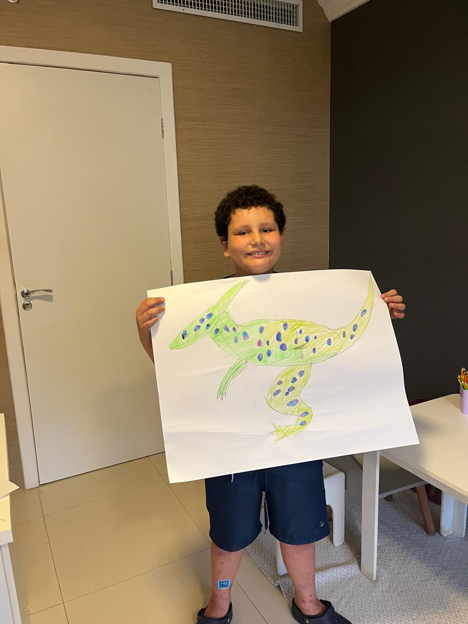 Menino sorridente segura um desenho detalhado de uma lagartixa verde com manchas coloridas.