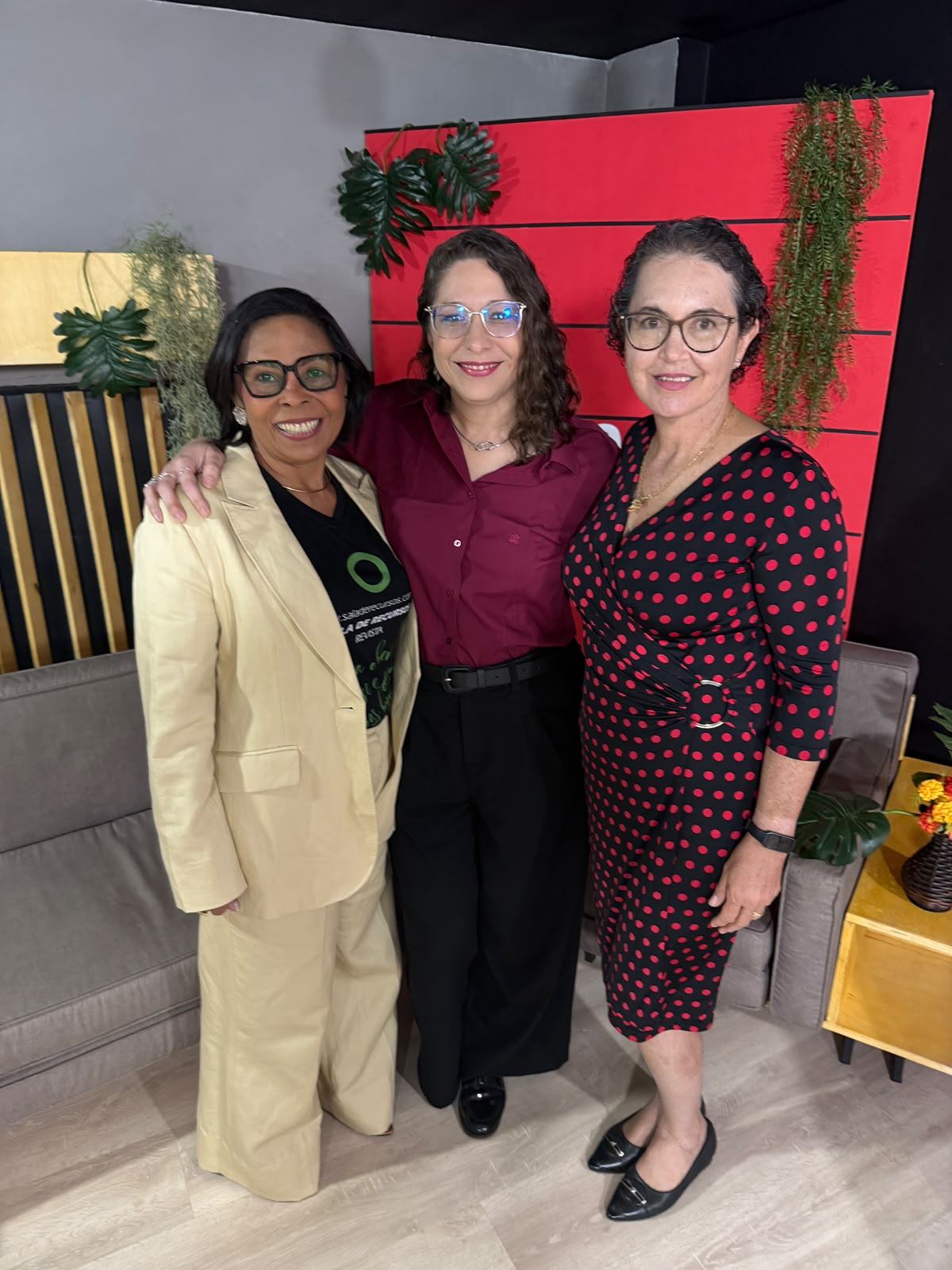 As três participantes lado a lado no estúdio da TV Comunitária de Brasília, diante de um painel vermelho e plantas. Da esquerda para a direita: Socorro Santos, de cabelo escuro, óculos de armação preta e blazer bege; Yesmin Correia Dias, de cabelo ondulado escuro, óculos de armação azulada e camisa vinho; e Fatima Cader-Nascimento, de cabelo curto e cacheado, óculos redondos e vestido preto com bolinhas vermelhas.